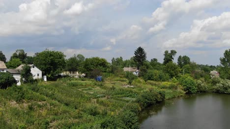 Niedrige-Luftaufnahme-Einer-Schroffen-Hütte-An-Einem-Fluss-Mit-Einem-Dock-Und-Häusern-Im-Hintergrund-An-Einem-Sonnigen-Tag