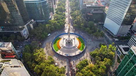 Aerial-of-Angel-of-Independence-victory-column-monument-on-a-roundabout-on-Paseo-de-la-Reforma-Avenue,-Day-of-the-Dead-parade-celebration-in-downtown-Mexico-street-on-sunny-daylight