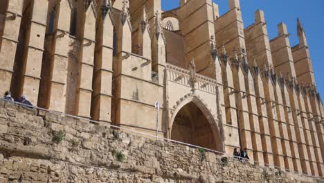 Tourist-looking-up-at-the-stunning-Cathedral-of-Mallorca's-exterior-wall,-soaking-in-the-historical-architecture-on-a-bright-sunny-day