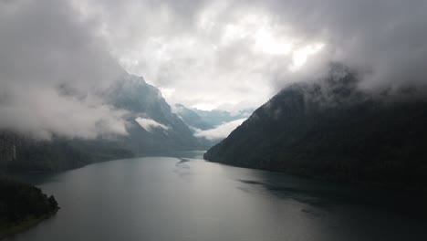 Drone-shot-of-a-Alpine-lake-after-rain-surrounded-by-high-mountains-with-low-clouds-in-Switzerland-Klontalersee