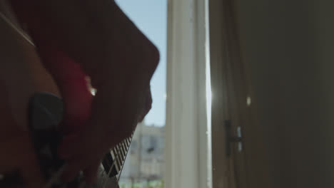 Beautiful-slow-motion-shot-of-hands-playing-electric-bass-in-a-room-as-sunlight-streams-in-through-the-balcony-backlit-in-front-of-the-camera