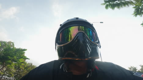 front-view-of-biker-riding-a-motorcycle-with-helmet-and-goggles-,-driving-in-nature-with-tall-tree