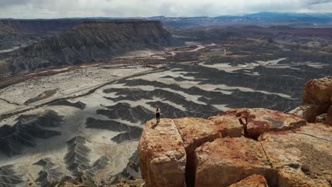 Toma-Aérea-De-Una-Joven-Caminando-Sobre-Un-Acantilado-Sobre-El-Abismo-Y-La-Inmensidad-Del-Paisaje-Desértico,-Utah,-EE.-UU.