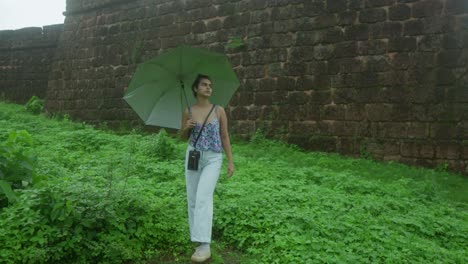 Young-woman-with-umbrella-exploring-historic-stone-wall-surrounded-by-lush-green-foliage