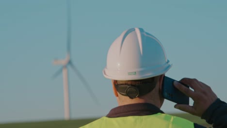 Engineer-in-hard-hat-and-safety-vest-talking-on-phone-near-wind-turbine-at-sunset