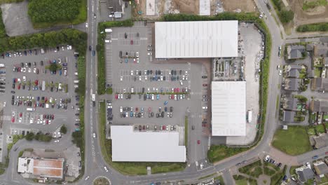 Drone-top-down-fly-out-of-a-UK-retail-park-and-carpark,-showing-the-surrounding-area,-including-roads,-green-spaces,-and-adjacent-buildings