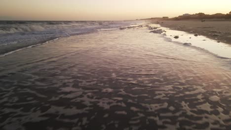 Flying-over-beach-water-at-sunset-then-transitioning-to-top-down-view-while-waves-roll-in