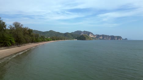 Krabi-Ao-Nang,-Sea-washing-a-tropical-beach-with-tourists-walking-on-the-sand-and-green-hills-in-the-background