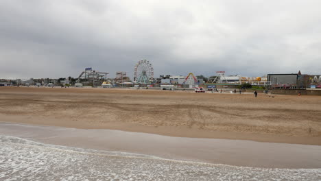 A-deserted-beach-stretches-towards-a-quiet-amusement-park-featuring-a-Ferris-wheel-and-roller-coaster-under-an-overcast-sky-at-skegness