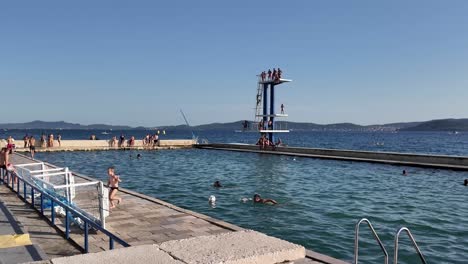 Sea-pool-and-beach-with-kids-playing-with-a-ball-and-jumping-board-in-the-background-in-Zadar,-Kolovare-beach-Croatia