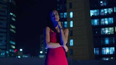 Una-Joven-Con-Un-Vestido-Rojo-Se-Encuentra-En-Un-Tejado-Por-La-Noche,-Con-Los-Edificios-De-La-Ciudad-Formando-Un-Telón-De-Fondo.