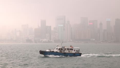 Patrulla-De-Barcos-De-La-Policía-Navegando-Por-El-Puerto-De-Victoria-En-Hong-Kong,-El-Horizonte-De-La-Ciudad-En-Segundo-Plano.