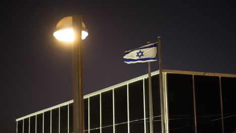 Die-Israelische-Flagge-Flattert-Im-Wind.-Die-Israelische-Flagge-In-Weiß-Und-Blau-Mit-Dem-Davidstern-Flattert-Nachts-Im-Wind-über-Dem-Dach-Eines-Gebäudes.