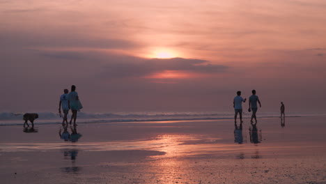 Parejas-Siluetas-Caminando-Sobre-El-Paseo-Marítimo-De-Arena-Húmeda-Y-Reflectante-En-El-Impresionante-Atardecer-De-Cielo-Púrpura-Rosa-En-Cámara-Lenta,-Playa-De-Jimbaran-En-Bali-Indonesia