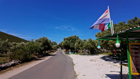 Pequeña-Tienda-Con-Banderas-Holandesas-A-Lo-Largo-De-La-Calle-Bordeada-De-Olivos-Cerca-De-Zakinthos-En-Grecia