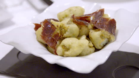 Primer-Plano-De-Una-Persona-Que-Utiliza-Un-Tenedor-Para-Coger-Un-Trozo-De-Un-Plato-Frito-Adornado-Con-Finas-Lonchas-De-Carne-Curada,-Resaltando-Su-Textura-Crujiente-Y-Su-Aspecto-Apetitoso.