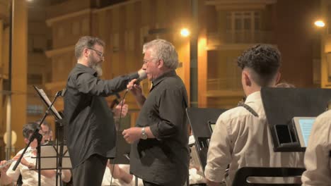 Man-in-black-shirt-singing-in-accompaniment-of-an-orchestra-at-an-evening-concert-with-conductor-behind-him-in-Sagunto