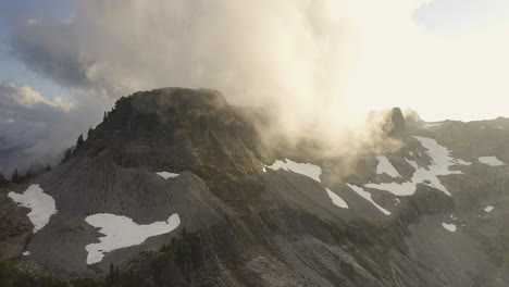 Drone-ascends-rocky-exposed-cliffs-near-Mt-Baker-in-PNW-with-golden-soft-light-diffused-between-clouds