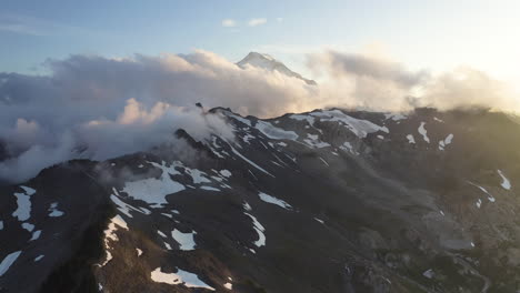 Aerial-pullback-at-sunset-of-Mount-Baker-golden-hour-glow-cast-along-snow-in-valley-with-clouds-rising-between-ridges