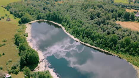 Aerial-view-of-lake-reflecting-a-cloudy-sky,-surrounded-by-dense-forest-and-open-fields-in-a-remote-countryside-setting