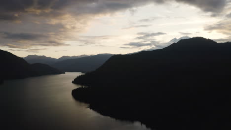 Silhouette-of-evergreen-forest-and-mountain-peaks-with-orange-glow-on-clouds,-aerial-descend