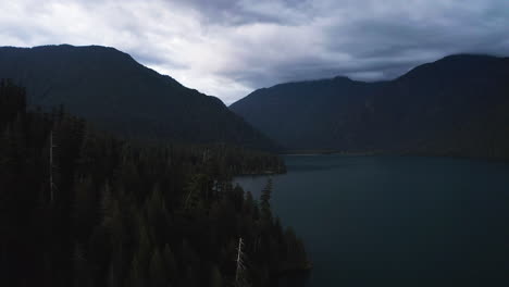 Silhouette-of-PNW-forest-above-alpine-lake-with-mountain-ridgelines-against-white-and-grey-clouds