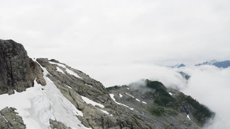 Glaciers-and-snow-fill-cracks-between-rocky-mountains-as-wispy-clouds-rise-above-evergreen-forest-and-drip-below,-aerial