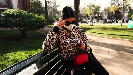 At-the-park-sitting-on-a-bench,-a-woman-texting-and-worried-on-her-smartphone-wearing-a-covid-mask,-dark-sunglasses,-long-black-hair,-an-animal-print-sweater,-black-pants-with-a-red-waist-leather-bag