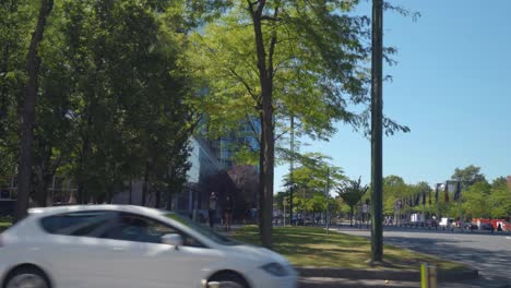 People-walking-across-urban-park-with-cars-passing-by-in-foreground-on-a-warm-sunny-day-with-clear-blue-skies-in-Brussels,-Belgium,-in-downtown-business-district