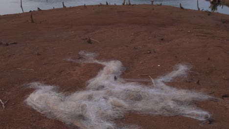 Tilting-down-shot-of-an-old-abandoned-white-fishing-net-on-the-beach-with-view-of-lake-and-mountain-range-on-the-background