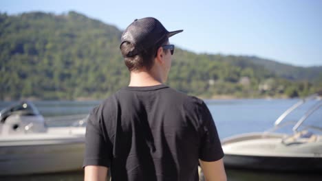 Un-Joven-Caminando-Sobre-Un-Pontón-En-Un-Lago,-Mirando-Lanchas-Rápidas-De-Arte-En-Un-Día-Soleado.