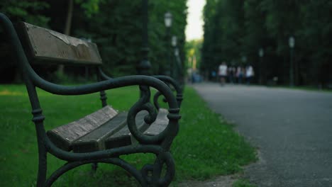 Banco-Del-Parque-Vacío-A-Lo-Largo-De-Un-Camino-Pavimentado-Con-Gente-En-El-Fondo-En-El-Parque-Maksimir