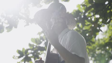 Man-in-straw-hat-playing-guitar-outdoors-under-tropical-trees,-Bali-vibe,-warm-sunlight