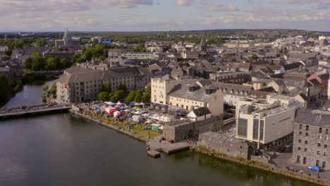 Aerial-rising-shot-of-bustling-Galway-city,-showcasing-the-market-at-Spanish-Arch-beside-the-River-Corrib