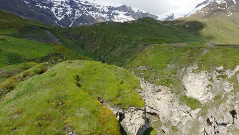 Aerial-orbit-of-hiker-on-grassy-slope-overlooking-ravine,-lake-and-mountains,-France