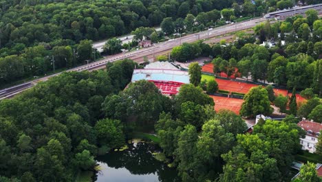 Estadio-De-Tenis-Con-Canchas-De-Arcilla-Roja,-Rodeado-Por-Un-Frondoso-Bosque-Verde-Y-Un-Pequeño-Lago.