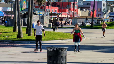 Couple-practices-rollerblading-as-man-jogs-without-shirt-on-at-Venice-Beach,-lots-of-people-exercising