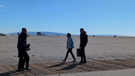 Couple-walks-along-boardwalk-at-Venice-Beach-on-sunny-day-in-winter-coats