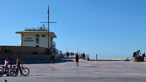 Familie-Geht-Mit-Dem-Fahrrad-Vor-Der-Küstenwache-Von-Manhattan-Beach-Spazieren,-Während-Touristen-Mit-Ihren-Hunden-Am-Pier-Entlang-Gehen