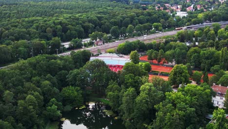 Tennis-stadium-with-red-clay-courts,-surrounded-by-a-lush-green-forest-and-a-small-lake