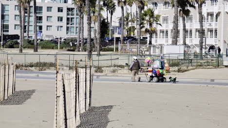 Venice-Beach-Schattennetze-Im-Sand-Aufgebaut,-Während-Menschen-Entlang-Des-Gehsteigs-Gehen-Und-Einen-Wagen-Mit-Taschen-Ziehen