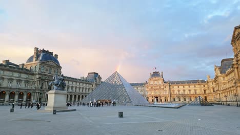 Rainbow-above-the-peak-of-the-Louvre-Glass-Pyramid-while-visitors-walk-towards-the-art-museum's-entrance-in-Paris,-France