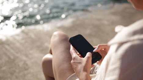 Frau-Sitzt-Am-Strand-Und-Tippt-Eine-Nachricht-Auf-Ihrem-Mobiltelefon