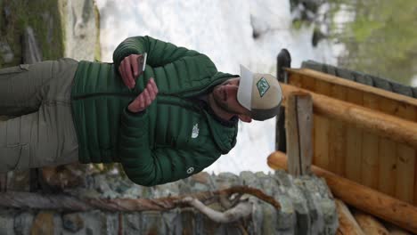 Man-in-a-green-jacket-and-cap-using-a-smartphone-outdoors,-near-a-wooden-structure-and-a-snowy-landscape