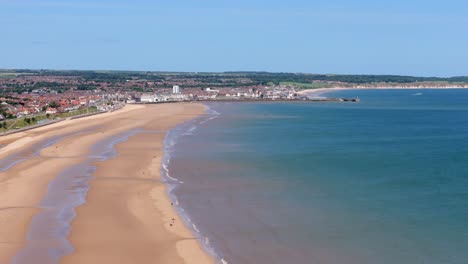 Imágenes-Aéreas-Sobre-La-Playa-De-Fraisthorp-Con-Bridlington-Al-Fondo-En-Verano-En-Un-Día-Caluroso-Y-Soleado,-Cielo-Azul-Y-Agua-Azul