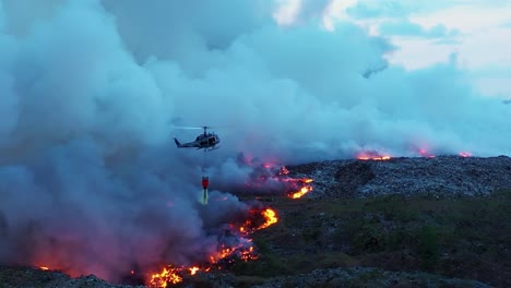 Aerial-drone-footage-of-a-UH-1H-HUEY-dropping-water-on-wildfires-in-the-Amazon-rainforest,-dense-smoke-rising-in-a-dark,-gloomy-evening,-in-Brazil,-South-America