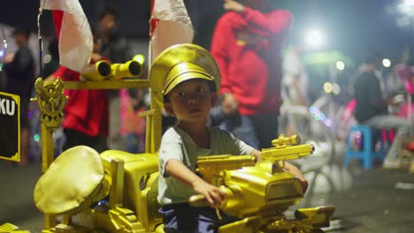 Un-Niño-Viaja-En-Un-Vehículo-De-Juguete-Dorado-En-Taman-Fatahillah-Durante-La-Noche-Rodeado-De-Gente-Y-Luces