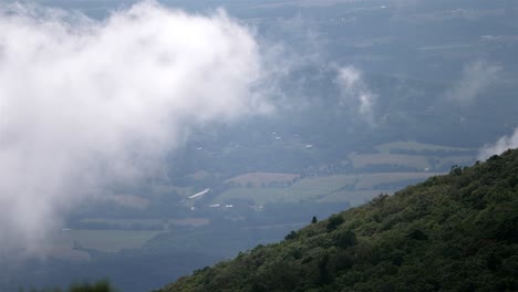 Gezoomte-Handaufnahmen-Von-Einem-Grünen-Hügel-Fangen-Einen-Atemberaubenden-Blick-Auf-Wolken-Ein,-Die-über-Entferntes-Ackerland-Ziehen