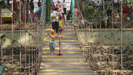 Indonesian-family-with-little-girl-leading-the-group-in-tricycle,-people-crossing-the-bridge