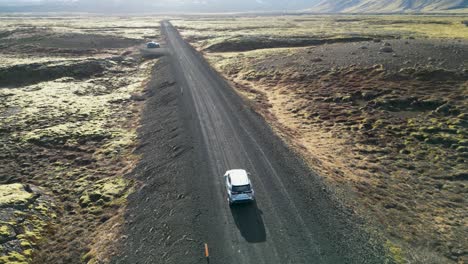 The-camera-shows-an-SUV-on-a-gravel-road-in-Iceland,-with-tall-mountains-and-green-mossy-terrain-in-the-background,-The-aerial-view-emphasizes-Iceland's-beautiful-landscape